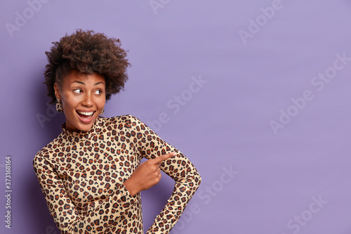 Happy feminine woman with curly hairstyle, points index finger to right side, discusses nice promo, gives direction to copy space, wears leoapard turtleneck, purple background, found what you search photo