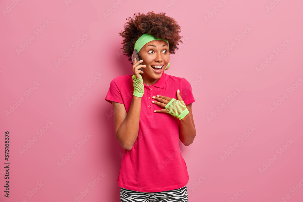 Overjoyed young Afro American woman feels excited, stares with surprised happy expression, holds breath, talks via smartphone, gasps wondered, receives great news, isolated on pink background