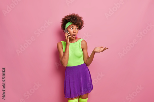 Confused dark skinned woman has shocked expression  tallks on mobile phone  stares in disbelief  raises palm  wears summer outfit  isolated on pink background. Communication. Human reaction.