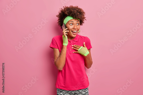 Overjoyed young Afro American woman feels excited, stares with surprised happy expression, holds breath, talks via smartphone, gasps wondered, receives great news, isolated on pink background