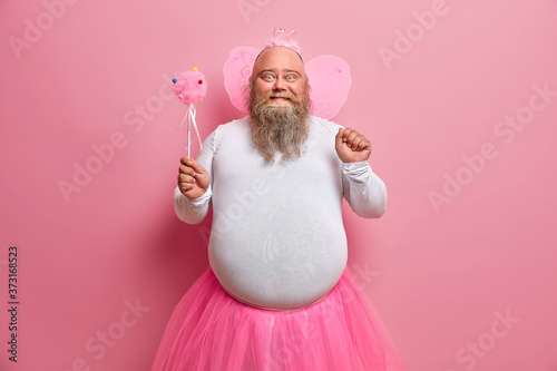 Murais de parede Positive plump man has fun on theme birthday party, feels like fairy who makes dreams come true, chills with children, has thick beard and fat belly, poses with magic wand and wings on back