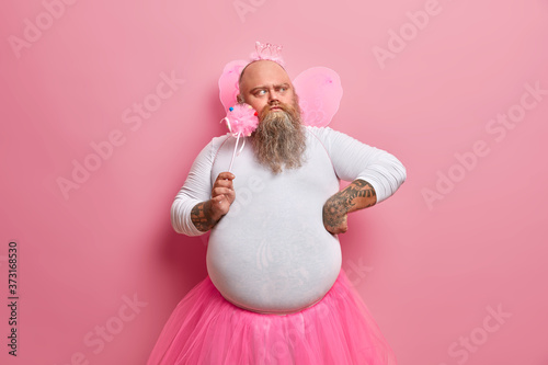 Fotografia Serious bearded man thinks how to entertain children on party, wears funny costume of princess or fairy, concentrated thoughtfully aside
