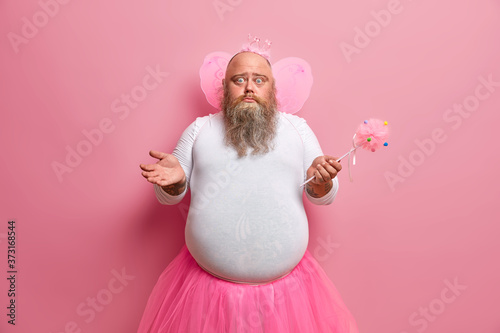 Canvas Print Big bearded man fairy with wings looks confused, spreads palms, organises costume party, poses with magic wand, has angelic expression, plays with daughter, wears princess crown