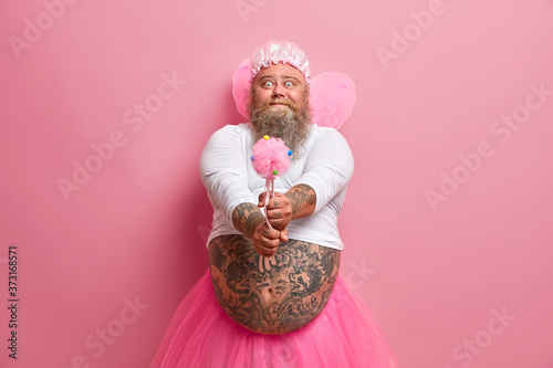 Photography Male performer or holiday animator works on party for little children, has funny expression, holds magic wand, dressed like fairy, isolated on pink background, wears fancy wings