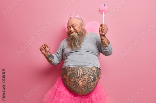Obraz na płótnie Overjoyed stout man in fairy costume dances carefree, has fun and foolishes around, organises holiday for children, plays princess, moves against pink background