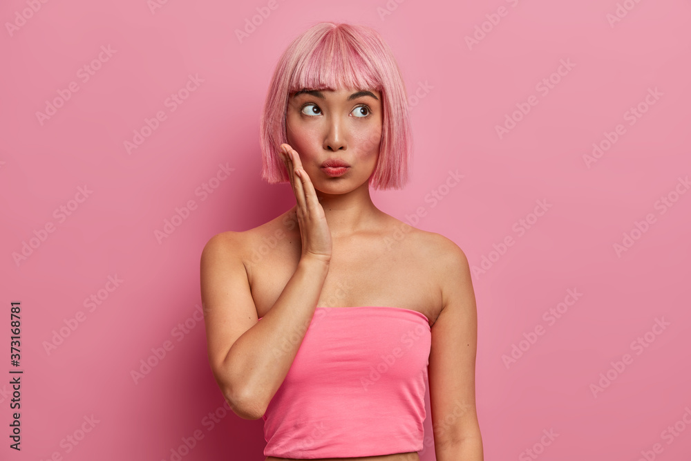 Pensive pink haired woman touches cheek, looks above, concentrated on something, dressed in casual tank top, shows bare shoulders, has trendy rosy hair, models in studio, thinks about offer.