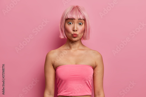 Women, facial expressions concept. Surprised young Asian woman keeps lips folded, looks with wonder at camera, wears rosy tank top, has pink hairstyle, hears something astonishing, stands indoor