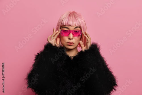 Gorgeous self confident serious woman wears trendy pink sunglasses, has rosy bob hair, dressed in fluffy warm black sweater, stands indoor, thinks about something. Women, fashion, style concept