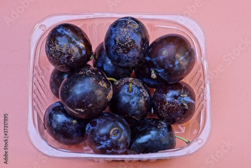 ripe blue plums in a transparent plastic box stands on a pink table