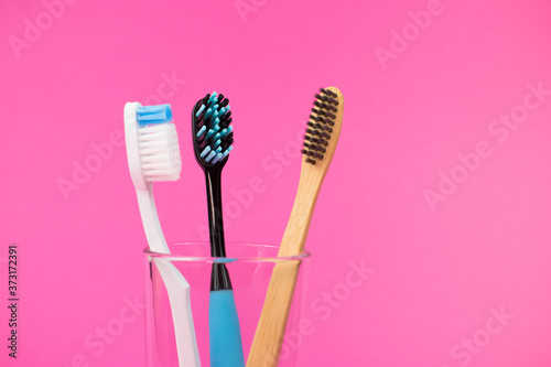 Ecological bamboo toothbrush with two ordinary brushes in a glass glass on a pink background with free space for an inscription.