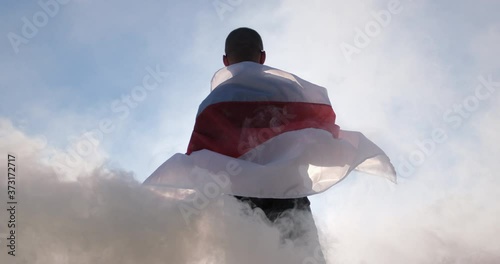 Belarus in a mask and sunglasses holds the flag of Belarus stretched in his hands in the backlight of the sun against a background of smoke. photo