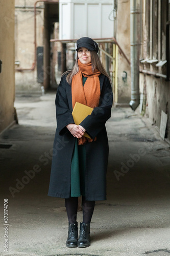 Long-haired girl student in a black coat with a vintage cap and a red scarf, posing with a book in a shabby quiet courtyard