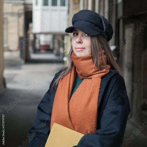 Long-haired girl student in a black coat with a vintage cap and a red scarf, posing with a book in a shabby quiet courtyard