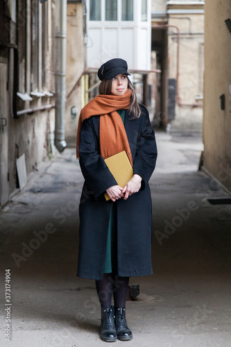 Long-haired girl student in a black coat with a vintage cap and a red scarf, posing with a book in a shabby quiet courtyard