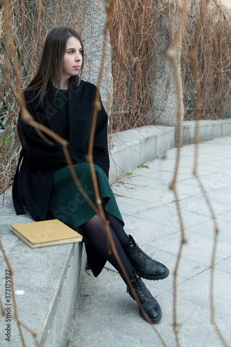 Brunette long haired girl sitting on a bench in a spring park near hedge without leaves