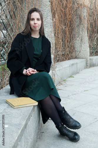 Brunette long haired girl sitting on a bench in a spring park near hedge without leaves