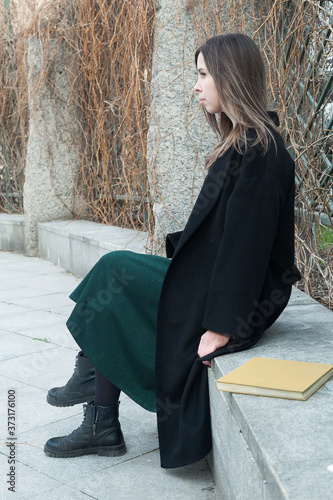 Brunette long haired girl sitting on a bench in a spring park near hedge without leaves