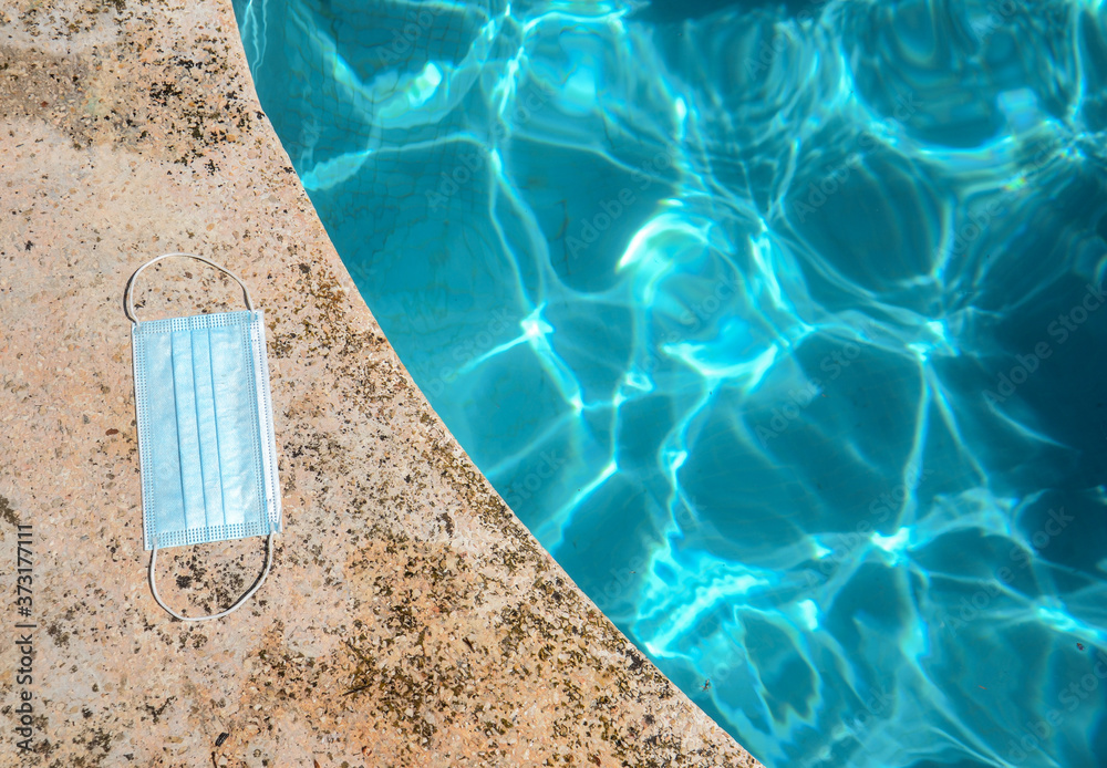 Blue face mask for protection against the Corona Virus (COVID-19), laying beside a swimming pool during coronavirus worldwide pandemic holidays Blue face mask for protection against the Corona Virus (COVID-19), laying beside a swimming pool during coronavirus worldwide pandemic holidays