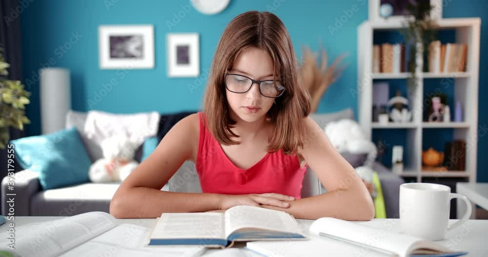 Front view of schoolgirl in eyeglasses sitting at table and reading interesting book. Concept of self education and kids activity during quarantine. Front view of schoolgirl in eyeglasses sitting at table and reading interesting book. Concept of self education and kids activity during quarantine.