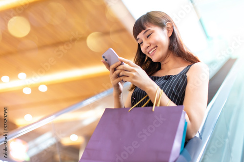 Asian young woman shopping online from smartphone. Happy woman with shopping bags enjoying in shopping. Girl holding colour paper bag and standing on escalator.time shopping .