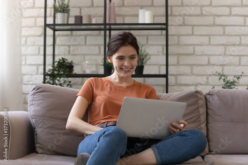 Smiling millennial Caucasian girl sit on couch in living room work study on laptop at home, happy young woman relax on sofa browse surf wireless internet, shopping online on gadget
