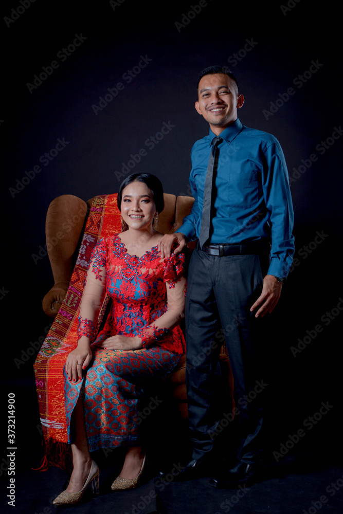 portrait of beautiful indonesian couple wearing traditional batak costume sit on brown couch isolated on black background.
