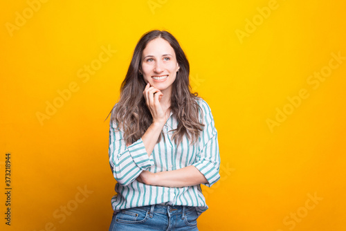 Young lady is holding hand near face, smiling and thinking about something over yellow background.