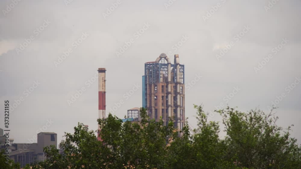 Panning shot of indian factory with red and white chimney and huge pipes for a distillation tower framed between trees and monsoon clouds