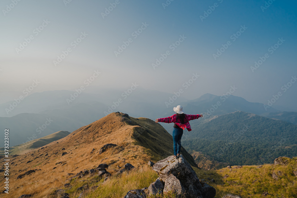 Obraz premium A woman with a red long-sleeved shirt and white hat stand and open arms on the stone at the ridge of Mulayit Taung, Myanmar. There are overlapping hills and golden dry grasslands. There is copy space.
