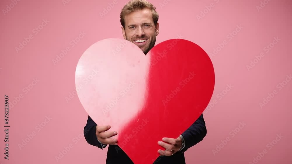 happy elegant businessman in navy blue suit smiling, holding and presenting big red heart on pink background happy elegant businessman in navy blue suit smiling, holding and presenting big red heart on pink background