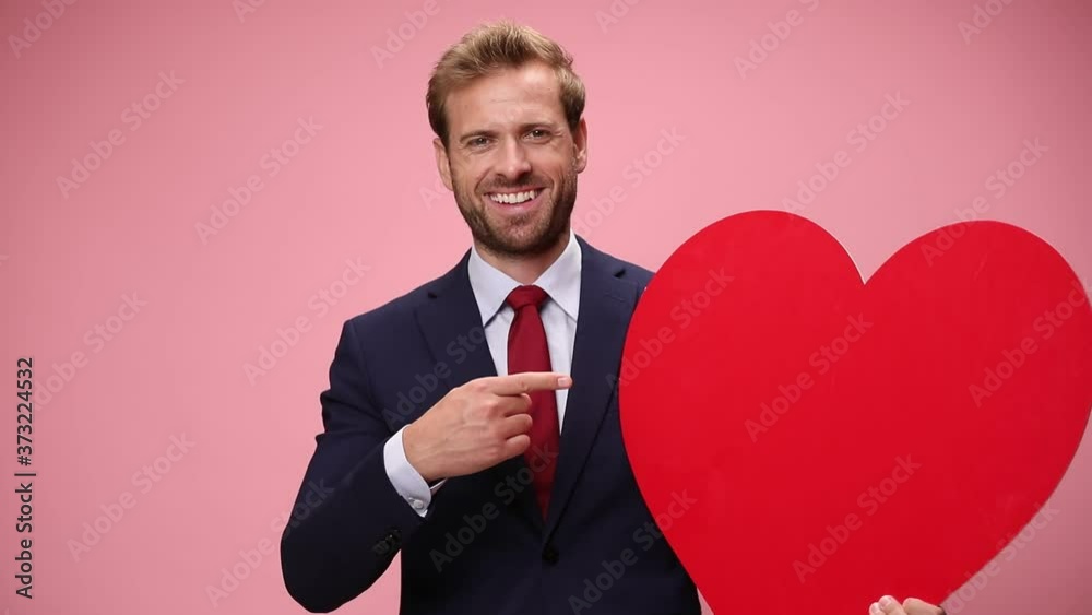 young elegant man holding big red heart, smiling and pointing fingers on pink background young elegant man holding big red heart, smiling and pointing fingers on pink background