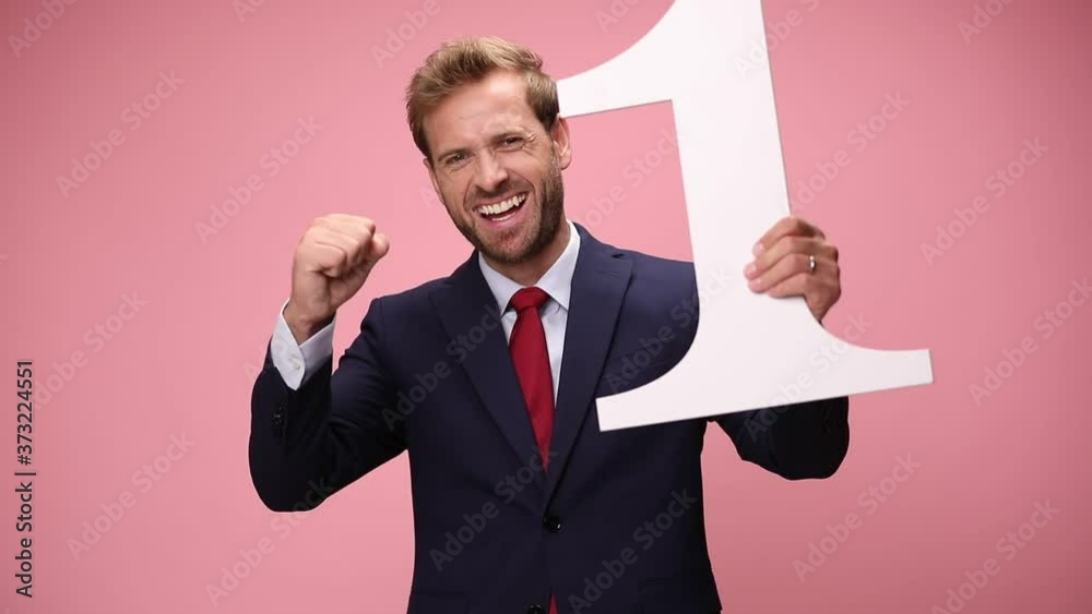 elegant young businessman in navy blue suit smiling, holding number one sign and fists in the air, celebrating victory on pink background elegant young businessman in navy blue suit smiling, holding number one sign and fists in the air, celebrating victory on pink background