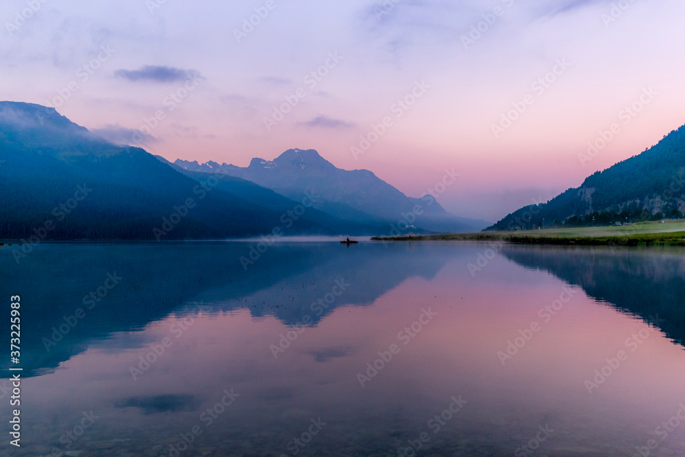 A fisherman rowing among the fog on the lake of Silvaplana in the Engadin valley at sunrise with mountains reflecting in the water A fisherman rowing among the fog on the lake of Silvaplana in the Engadin valley at sunrise with mountains reflecting in the water