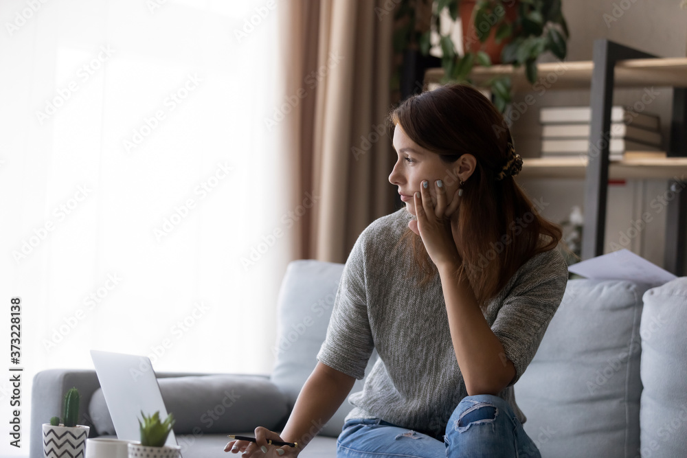 Fototapeta premium Pensive young woman manage household finances at home, look in distance thinking pondering of financial problem solution, thoughtful female sit on couch work on laptop decide or plan, lost on thoughts