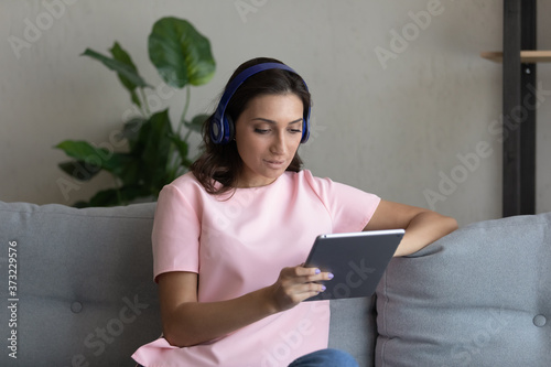 Young Arabic indian woman in modern wireless headphones sit relax on couch in living room watch video on tablet, arab mixed race female in earphones browse surf internet on pad, using gadget at home