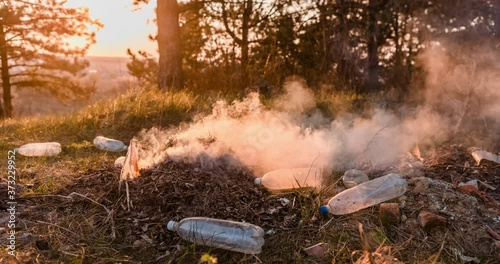 Time-lapse of a bunch of burning garbage left in the middle of a forest, during sunset, shot with a camera that's slowly moving to the left.