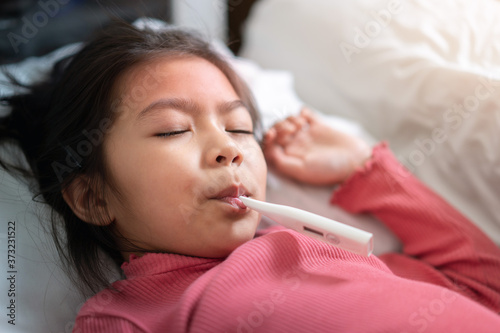 Asian girl with digital thermometer in her mouth on bed at morning time, Sick child with high fever Selective focus, Healthy and infection concept