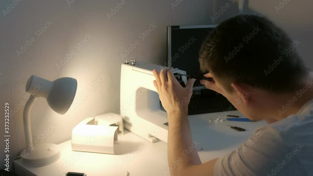 Young repairman master is repairing sewing machine screwing the bolt sitting at table, side view. Unscrew body of sewing machine using screwdriver. Man is testing and disassembles the sewing machine.