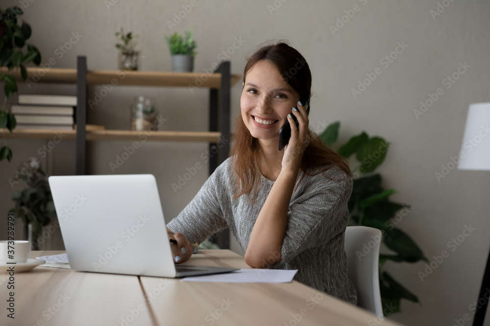 Portrait of smiling young Caucasian businesswoman sit at desk at home work on laptop talk on cellphone, happy woman have mobile online call or conversation on smartphone, technology concept Portrait of smiling young Caucasian businesswoman sit at desk at home work on laptop talk on cellphone, happy woman have mobile online call or conversation on smartphone, technology concept