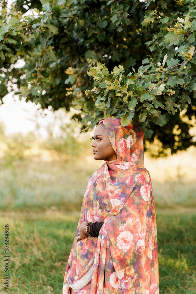 Muslim black woman African ethnicity weared traditional colorful hijab smiles and looks left side under green tree. Arabic islam religion clothes. Pretty black girl smiles Muslim black woman African ethnicity weared traditional colorful hijab smiles and looks left side under green tree. Arabic islam religion clothes. Pretty black girl smiles