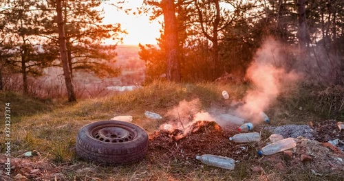 Time-lapse of a heap of burning garbage left in the middle of a green forest, during sunset, shot with a camera that's slowly moving to the right.