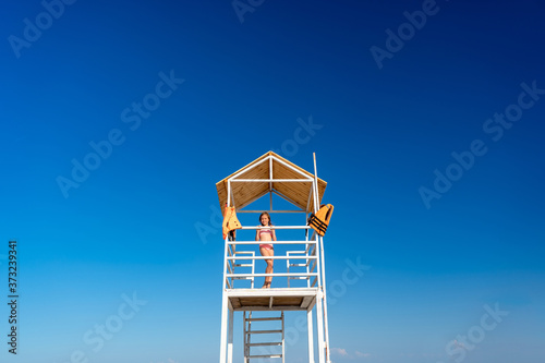 teenage girl stands on lifeguard tower on beach against cloudless sky.