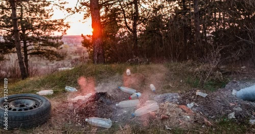 Time-lapse of a pile of burned garbage left in the middle of a forest, during sunset, shot with a camera that's slowly moving to the left.