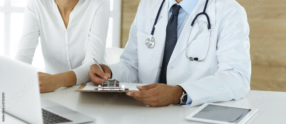 Doctor and patient discussing medical exam results while sitting at the desk in clinic, close-up. Male physician using clipboard for filling up medication history record of young woman Doctor and patient discussing medical exam results while sitting at the desk in clinic, close-up. Male physician using clipboard for filling up medication history record of young woman