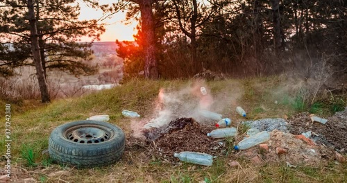 Time-lapse of a pile of burned garbage left in the middle of a forest, during sunset, shot with a camera that's slowly moving to the right.