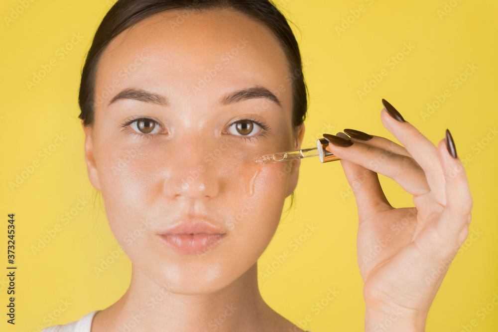 Fototapeta premium A beautiful young girl applies a serum to her face with a pipette. Cosmetology, skin care. Yellow background