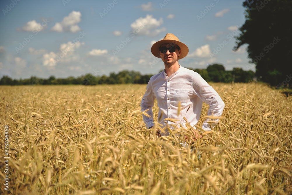 Young smiling man in wheat field on sun looks at camera. Positive young farmer in hat in middle of the field. Farmer inspecting ripe wheat crop development in field, adult male agronomist. Young smiling man in wheat field on sun looks at camera. Positive young farmer in hat in middle of the field. Farmer inspecting ripe wheat crop development in field, adult male agronomist.