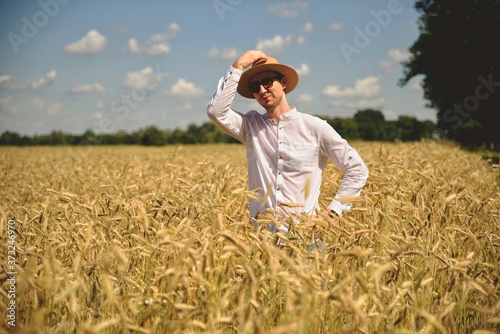 Young smiling man in wheat field on sun looks at camera. Positive young farmer in hat in middle of the field. Farmer inspecting ripe wheat crop development in field, adult male agronomist.