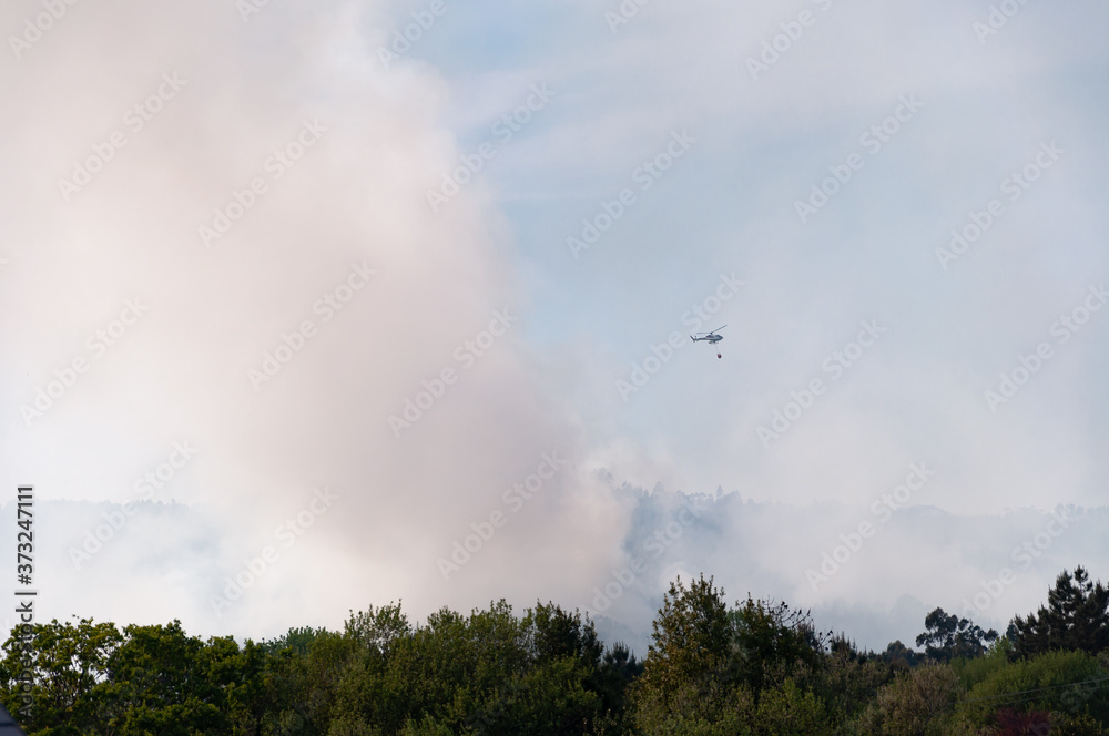 Helicopter firefighter flying over smoke fire in forest against blue and cloudy sky. 2017 Summer forest fires in Galicia Helicopter firefighter flying over smoke fire in forest against blue and cloudy sky. 2017 Summer forest fires in Galicia