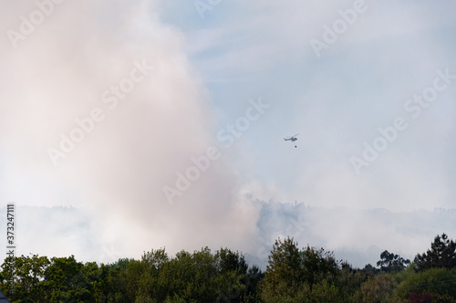 Helicopter firefighter flying over smoke fire in forest against blue and cloudy sky. 2017 Summer forest fires in Galicia